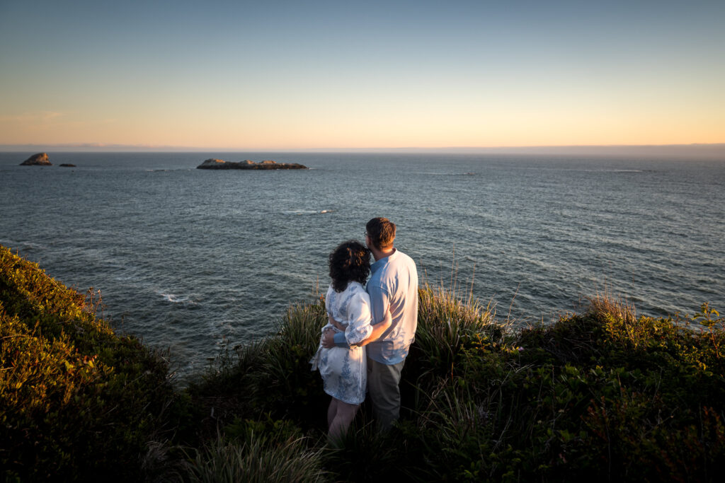 A couple standing on a coastal cliff, embracing as they look out over the ocean during golden hour, with small islands in the distance.