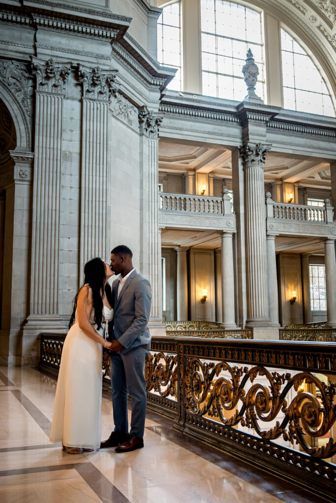Interracial couple sharing an intimate moment at San Francisco City Hall during their elopement photoshoot, standing close under natural light in the grand marble interior with elegant architecture and gold railings.