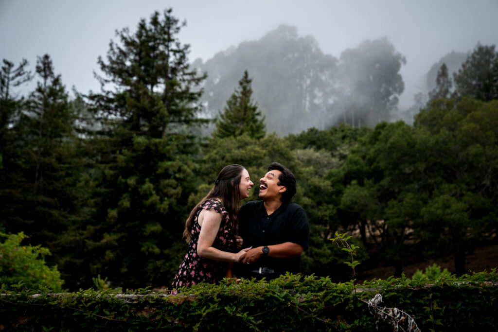 Couple laughing together on a stone wall with foggy forest at Joaquin Miller Park in Oakland