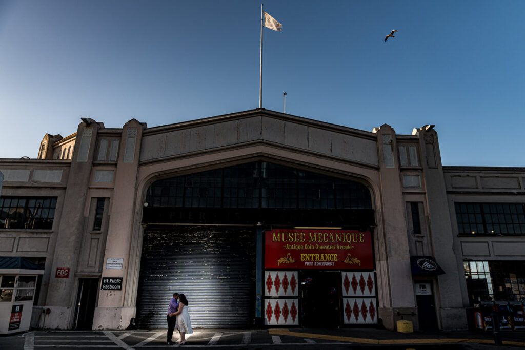 Couple embracing in front of Musée Mécanique at Fisherman’s Wharf with dramatic evening light.