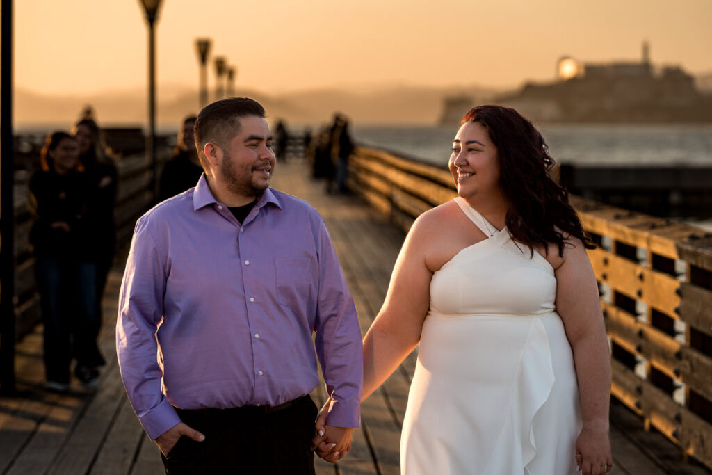 Couple holding hands and smiling at each other while walking along a pier at sunset with Alcatraz in the background.