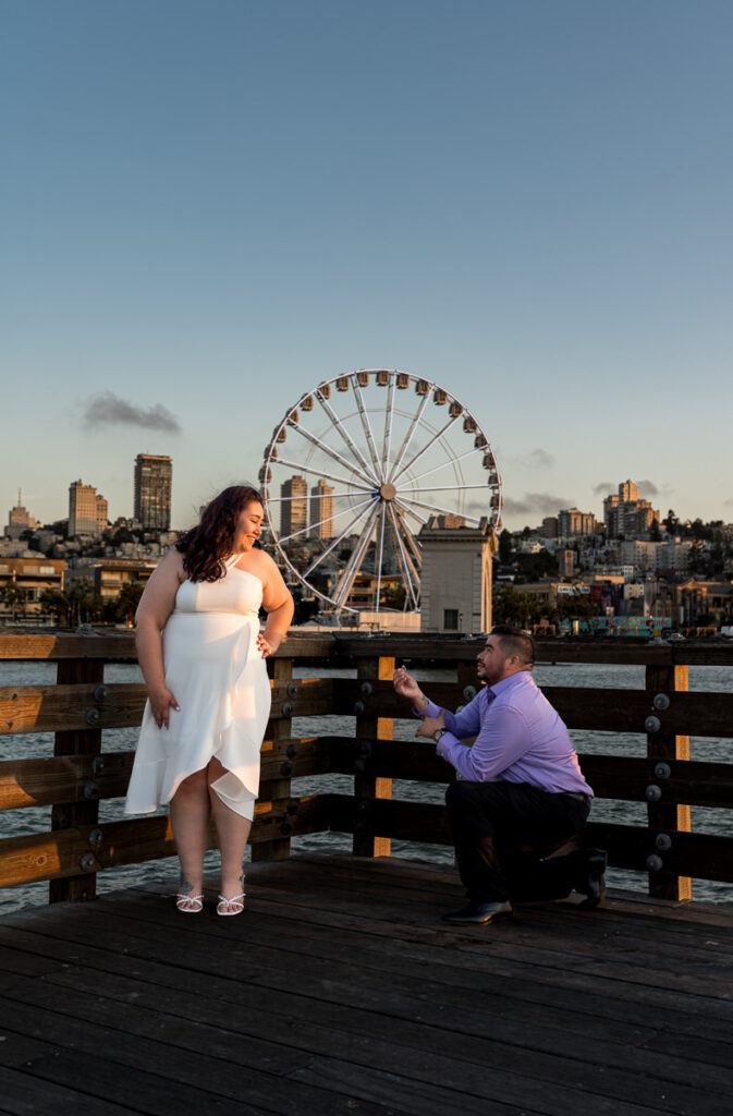 Man proposing to his partner on a pier with the SkyStar Wheel and San Francisco skyline in the background.