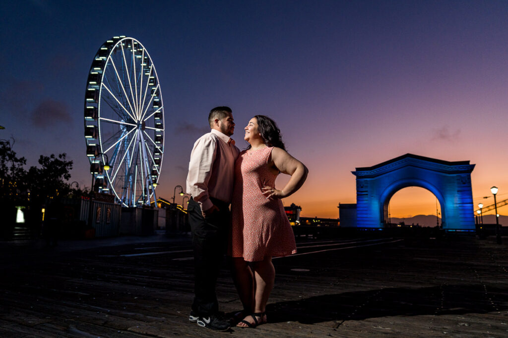 Couple standing close at Fisherman’s Wharf during blue hour, with SkyStar Wheel and Ferry Arch glowing in the background.