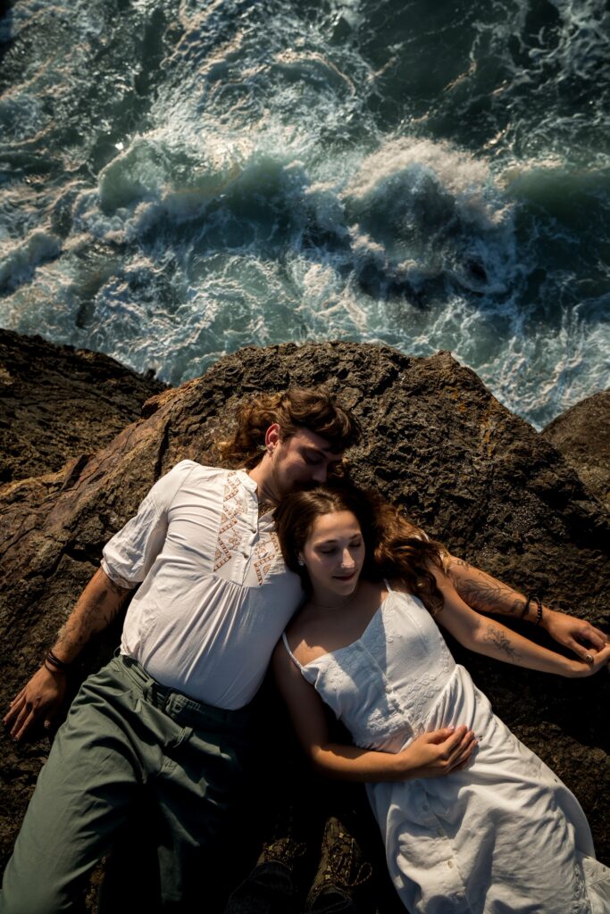 Couple lying on dark rocks in white outfits above crashing teal ocean waves at Elk Head Trail