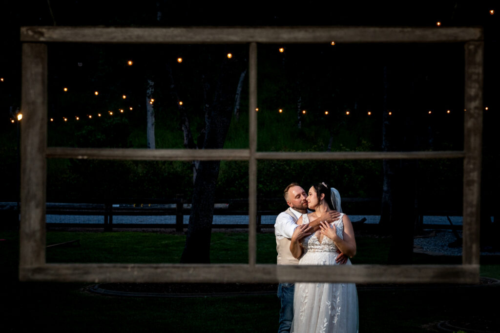 Bride and groom embracing at dusk, framed through a wooden window frame at TBS Ranch