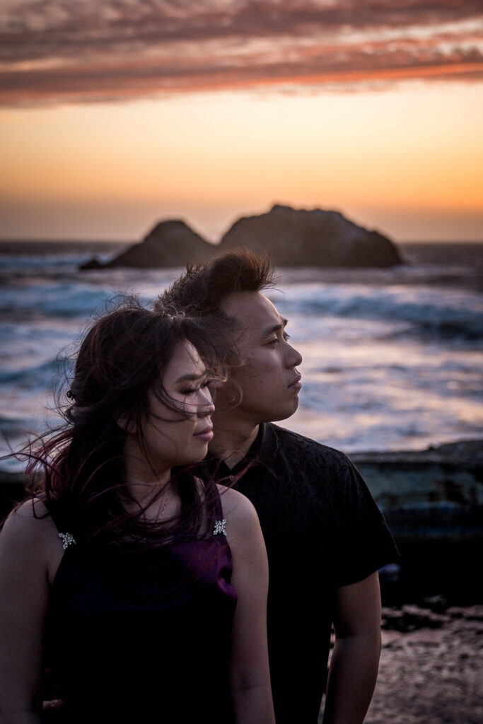 Dana and Kevin standing close with wind-blown hair against the ocean at sunset at the Sutro Baths