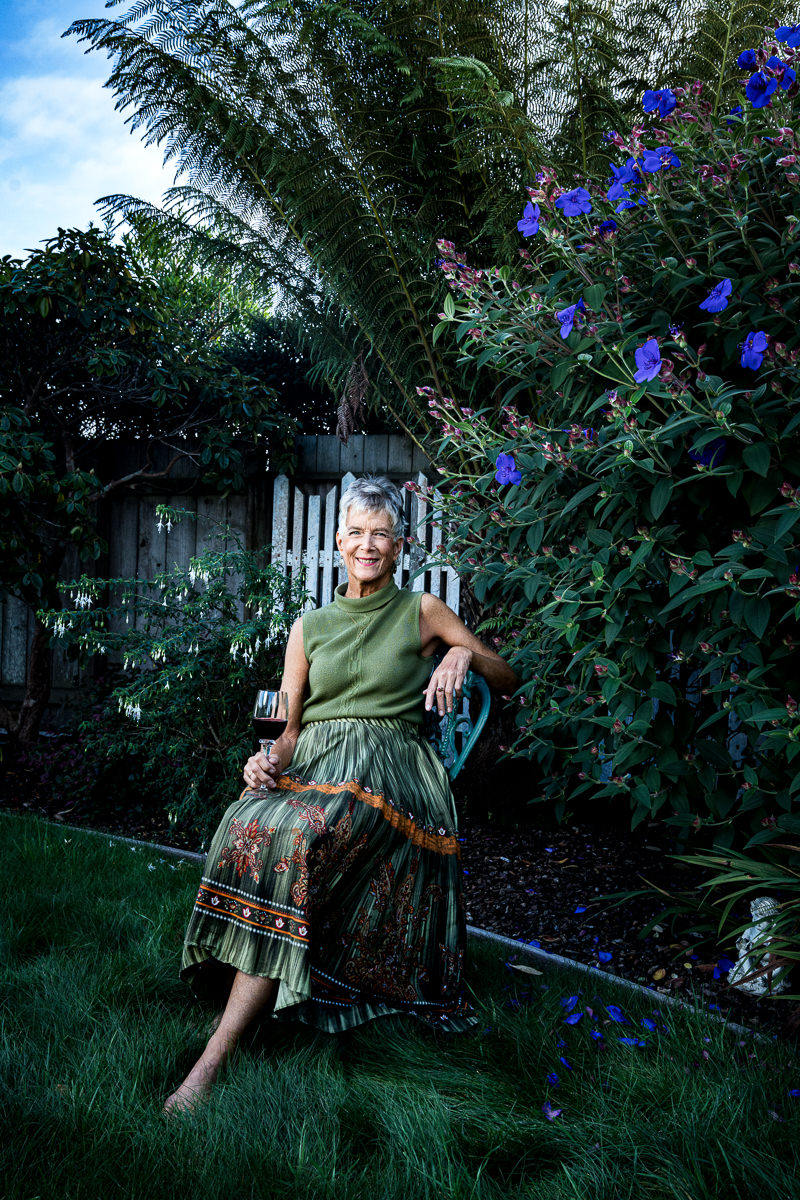 Portrait of Carol Clymo, a vibrant personality and long-time resident of Humboldt County. She sits in her backyard surrounded by lush greenery, reflecting her love for nature. The image captures her warmth and connection to the community, telling a story of a life well-lived in the heart of Humboldt.