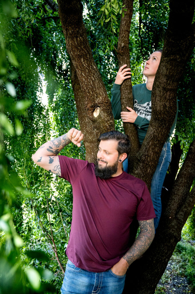 Emily rolls her eyes playfully as Dakota strikes a humorous pose with his arm up, both nestled among the branches of a lush tree in Hydesville, capturing a moment of light-hearted fun in their engagement session, photographed by Dan Berger Photography.