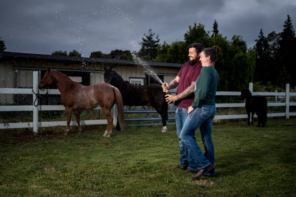 Emily and Dakota joyfully popping champagne in her dad's backyard in Hydesville, with the spray catching the light against a dramatic sky, celebrating their engagement in a playful moment by her horses, captured by Dan Berger Photography.