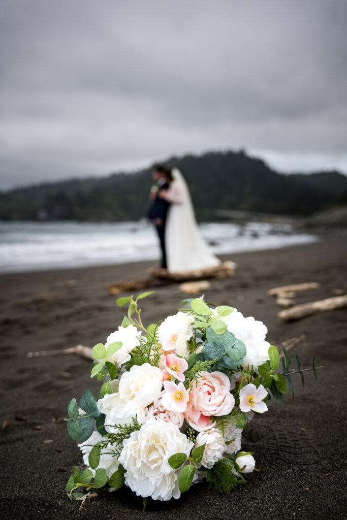 A close-up of a vibrant wedding bouquet resting on the sandy Wilson Creek Beach, with the bride and groom softly blurred in the background, capturing a romantic and dreamy coastal wedding moment.