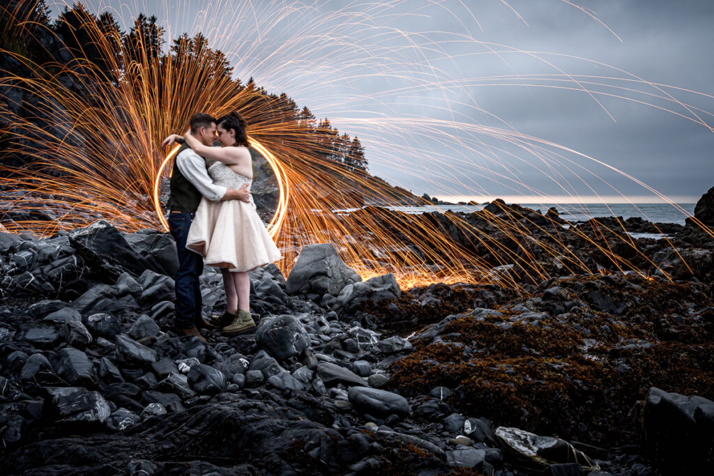 Bride and groom embracing with dazzling sparks behind them on Agate Beach at Sue-Meg State Park in Trinidad, framed by the rugged coastline and the thunderous waves of the Pacific Ocean.