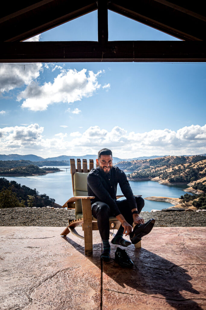 Groom sits on a rustic wooden chair at Airola Road Vineyard in Murphys, CA, putting on his shoes with a breathtaking view of New Melones Reservoir and the rolling Sierra Foothills behind him.