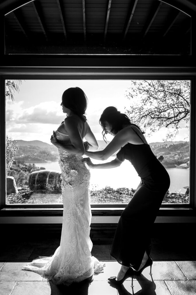 Black and white photo of the bride getting ready at Airola Road Vineyard, Murphys, CA, as her bridesmaid helps her into her dress in front of a large window overlooking the vineyards and mountains.