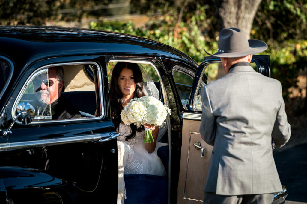 Bride steps out of a vintage black car at Airola Road Vineyard in Murphys, CA, holding her bouquet as she prepares to walk down the aisle for her vineyard wedding.