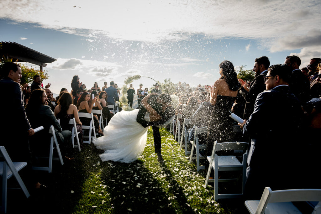 Bride and groom share a dramatic kiss at the end of the aisle as wedding guests celebrate with confetti cannons at Airola Road Vineyard in Murphys, CA.