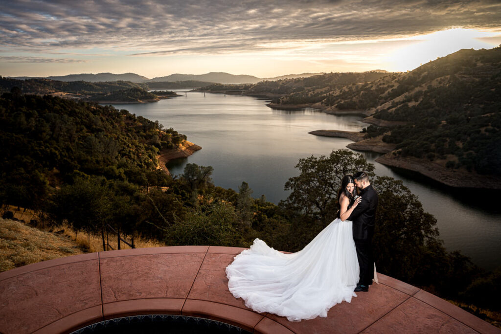 Bride and groom stand together on a scenic overlook at Airola Road Vineyard in Murphys, CA, with a breathtaking view of New Melones Reservoir and the Sierra Foothills at sunset.