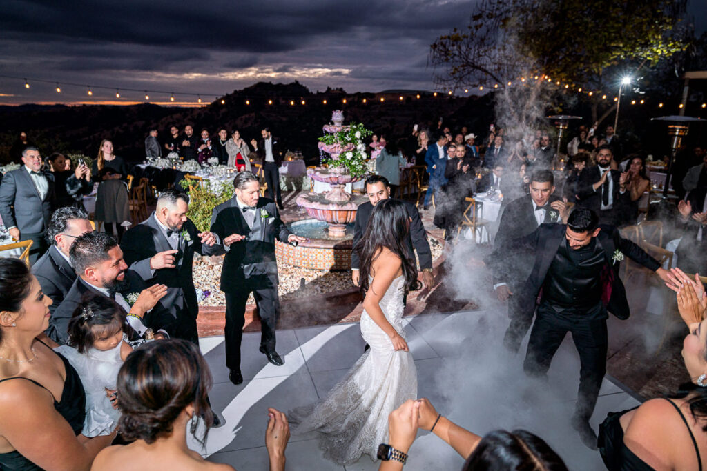 Bride and groom dance at Airola Road Vineyard in Murphys, CA, surrounded by guests as fog effects fill the dance floor. The overcast night sky and remnants of a dramatic sunset create a moody and energetic reception atmosphere.