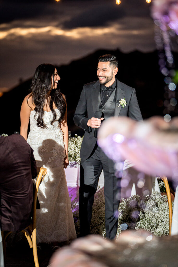 Bride and groom laugh together during their wedding toast at Airola Road Vineyard in Murphys, California, with a stunning twilight sky and floral-decorated tables in the background.